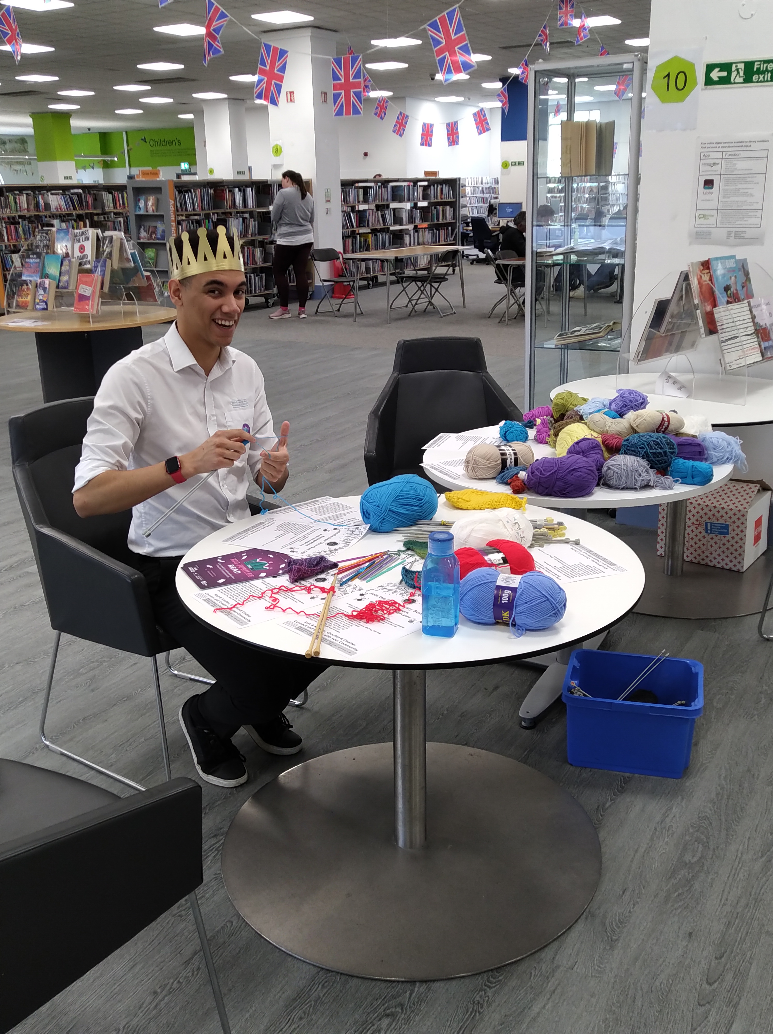 Smiling young man knitting in staff uniform sat at one of two tables with piles of yarn, knitting needles and pieces of paper