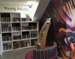 Colourful sofa, large white bookcase stacked with books and another smaller curved bookcase. The area is labelled 'Young Adults' and there is a large image of an eye on the right wall.